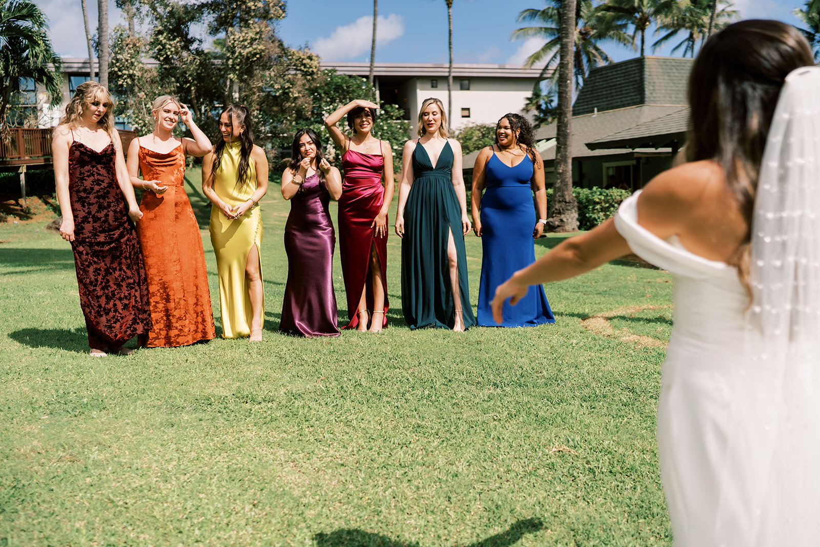 A bride in white gestures towards a group of smiling bridesmaids in colorful dresses standing on a lawn in Kauai