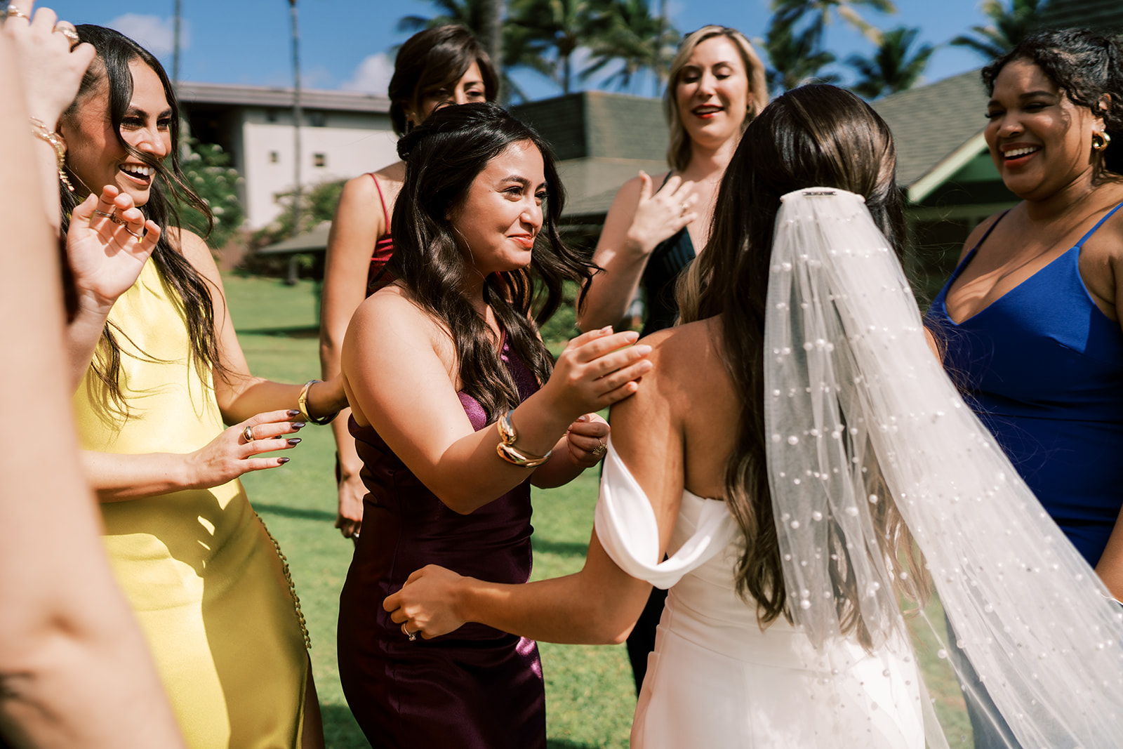 A bride in a white dress with a veil receiving congratulations from a group of smiling women in colorful dresses