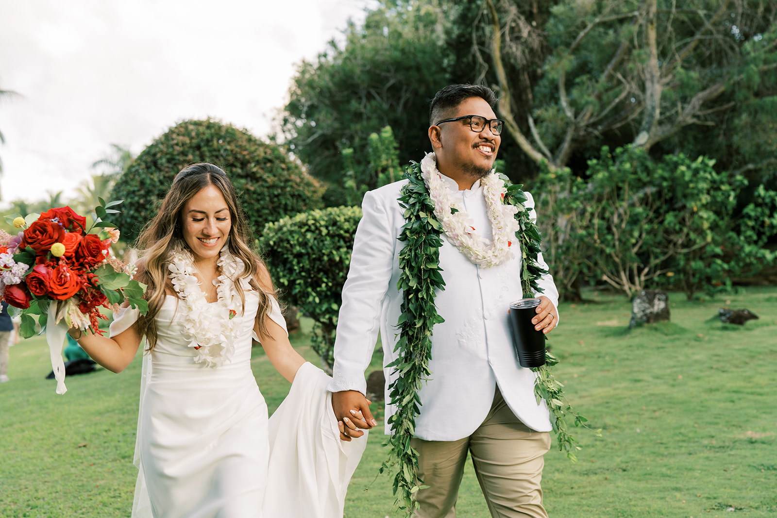 Bride and groom holding hands and walking on a grassy field, both wearing leis and smiling during their Hawaiian Wedding