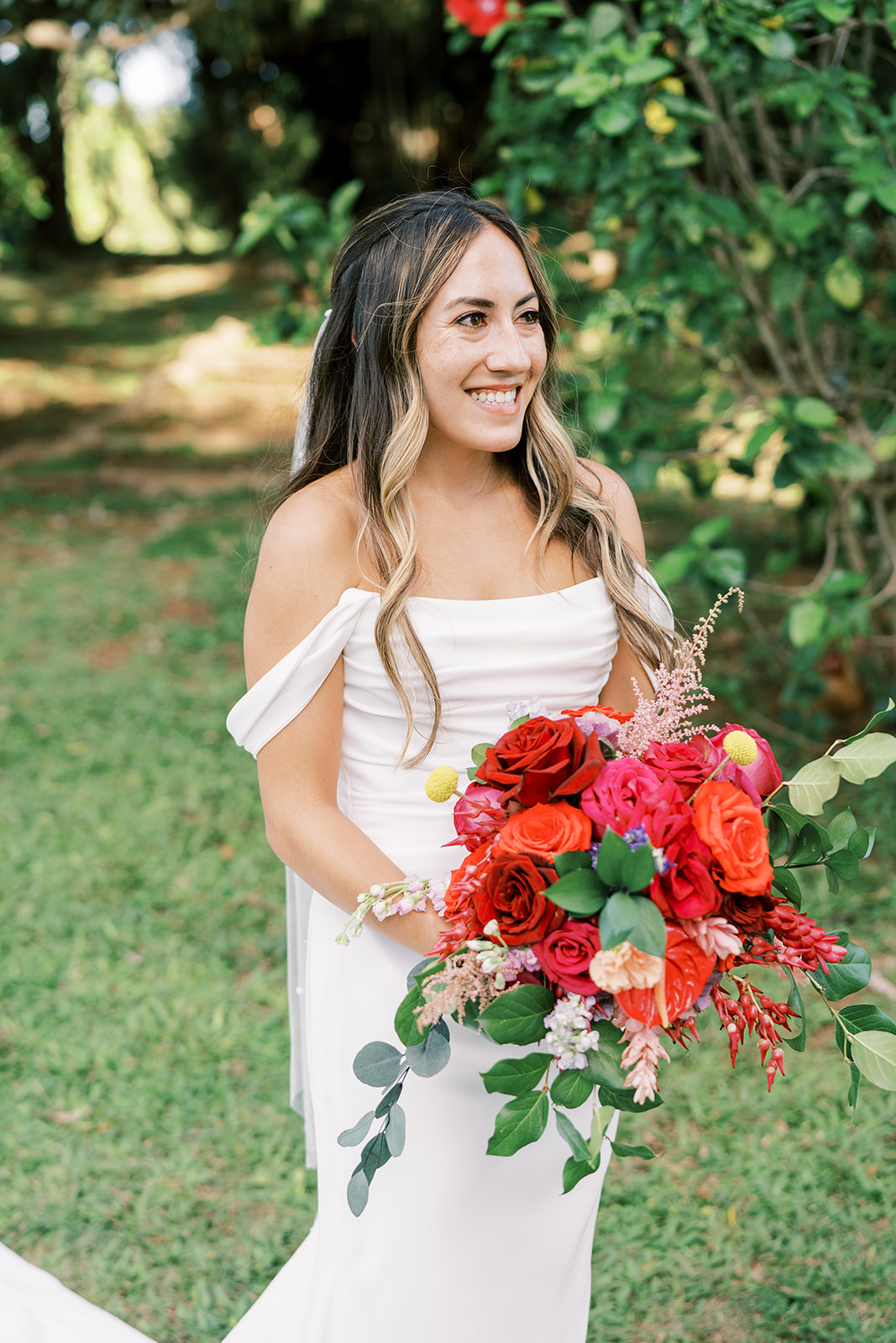 A smiling bride holding a bouquet of red flowers outdoors during her Hawaiian Wedding in Kauai