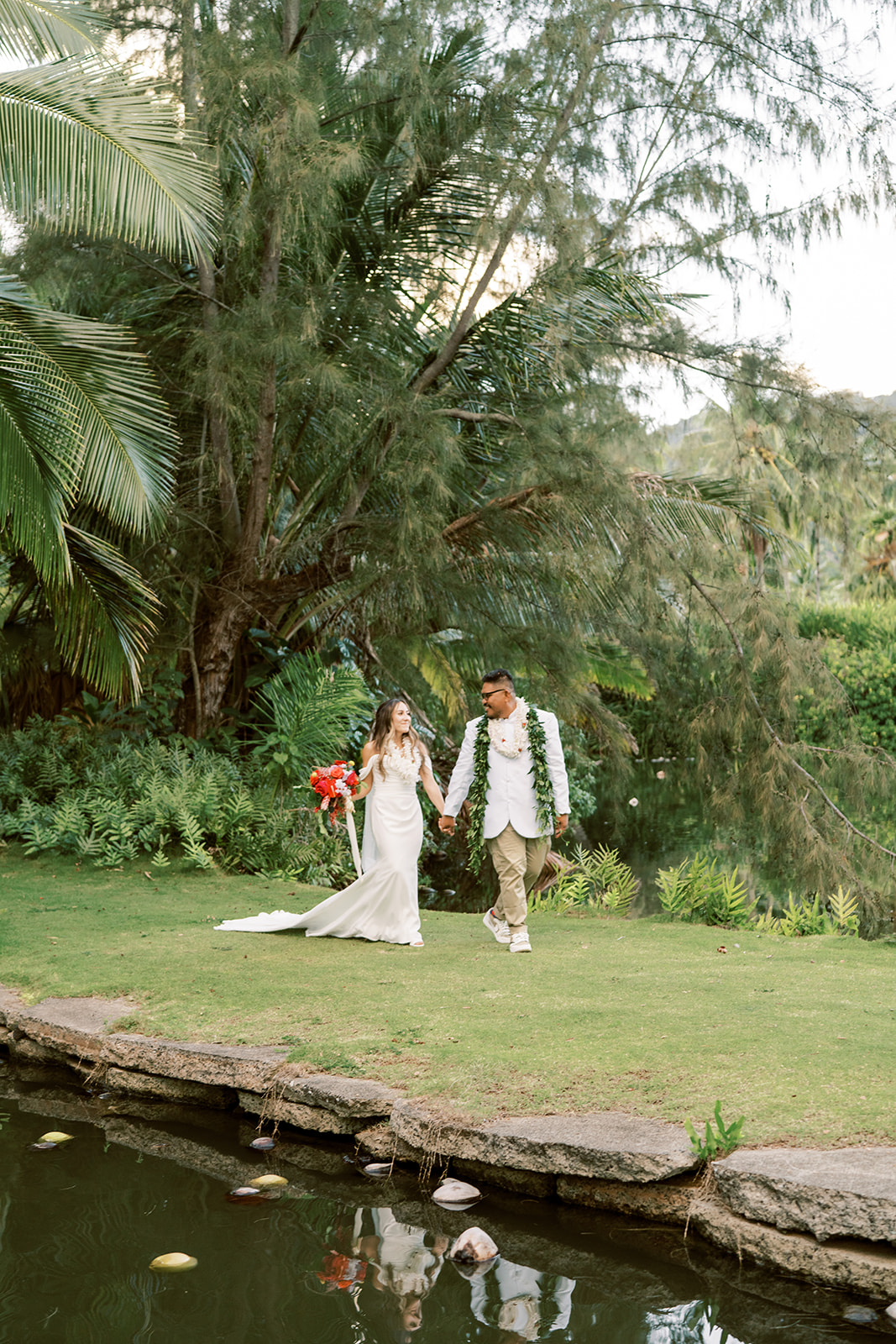 A couple in wedding attire holding hands and walking beside a pond in a lush garden setting of their Hawaiian Wedding