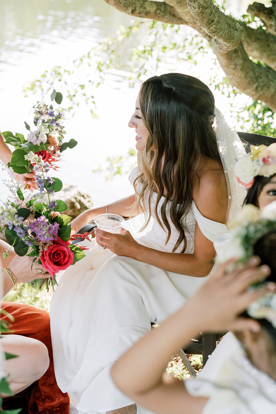 A bride sitting outdoors, laughing and holding a drink, with a colorful bouquet nearby Hawaiian Wedding in Kauai