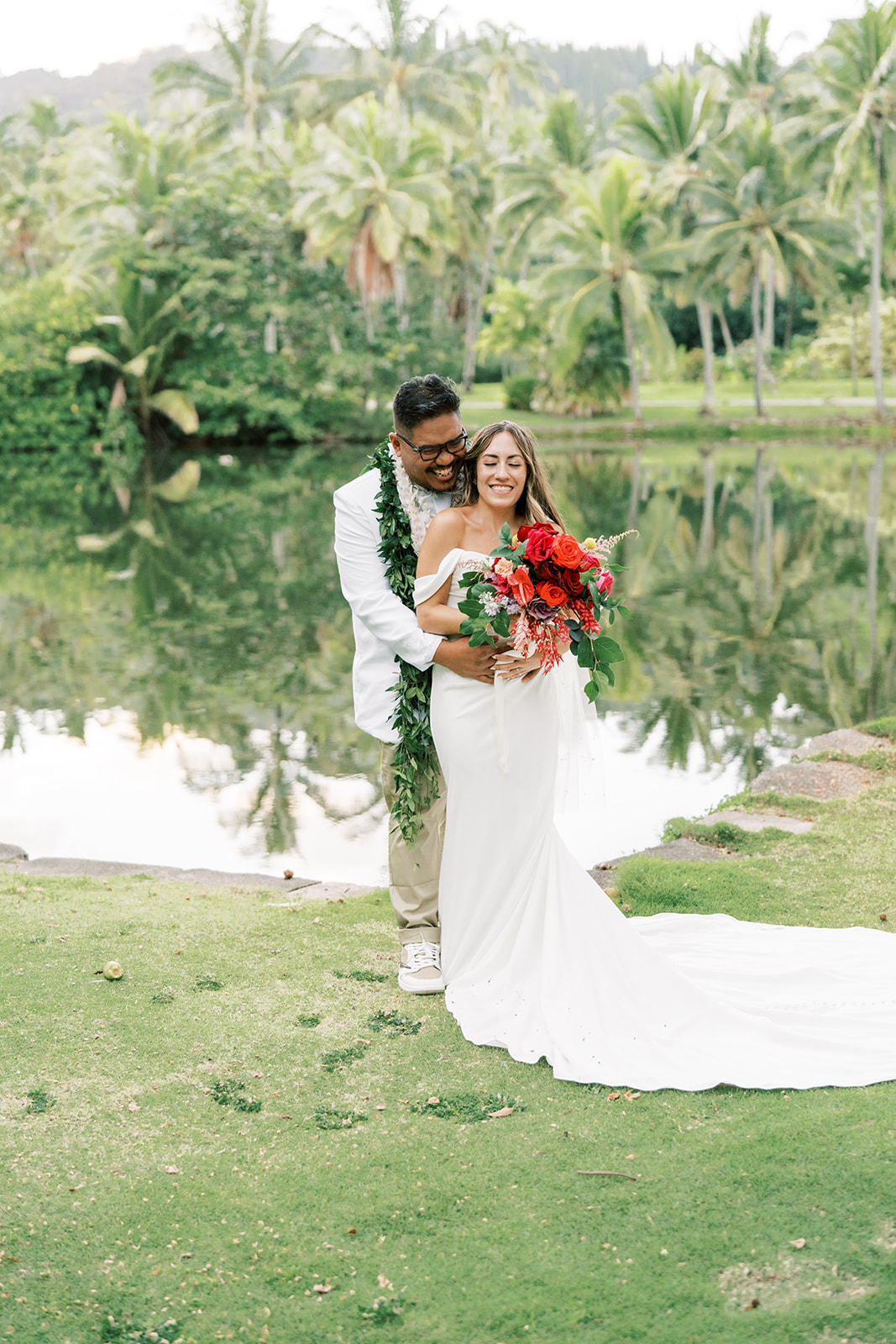 A joyful couple in wedding attire embracing beside a pond with lush greenery in the background.