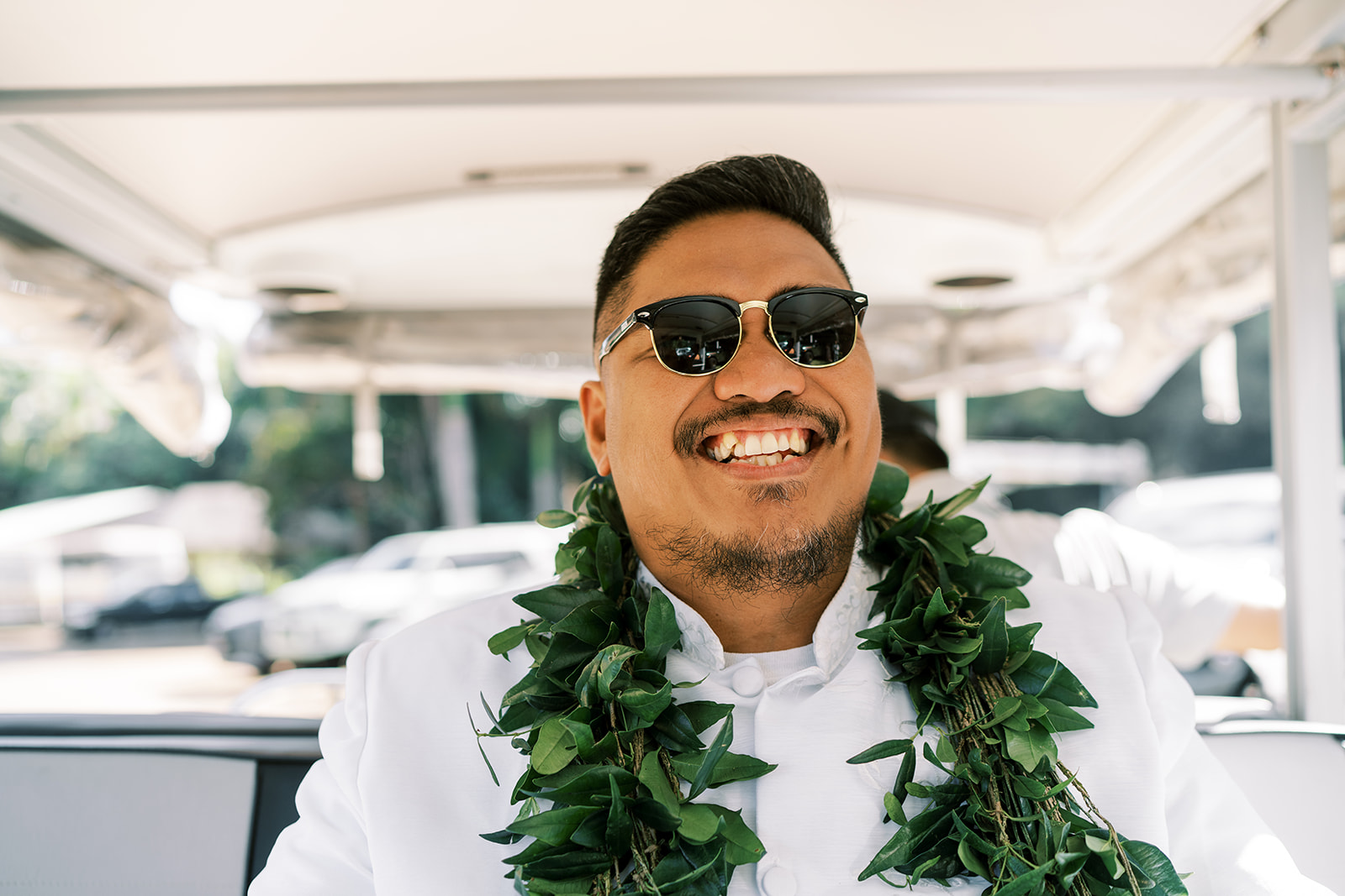 Man wearing sunglasses and a leaf lei smiling while riding  a cart during his Hawaiian  Wedding