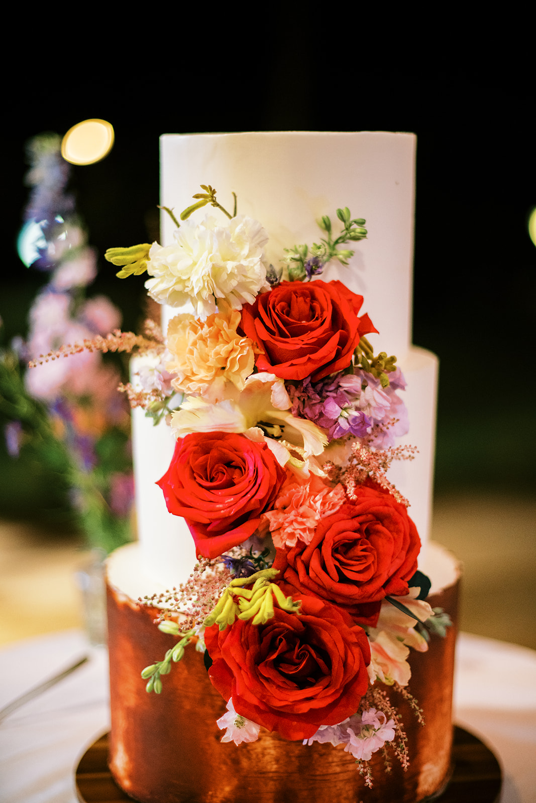 Three-tiered wedding cake adorned with red roses and assorted flowers captured by Megan Moura Wedding Photographer