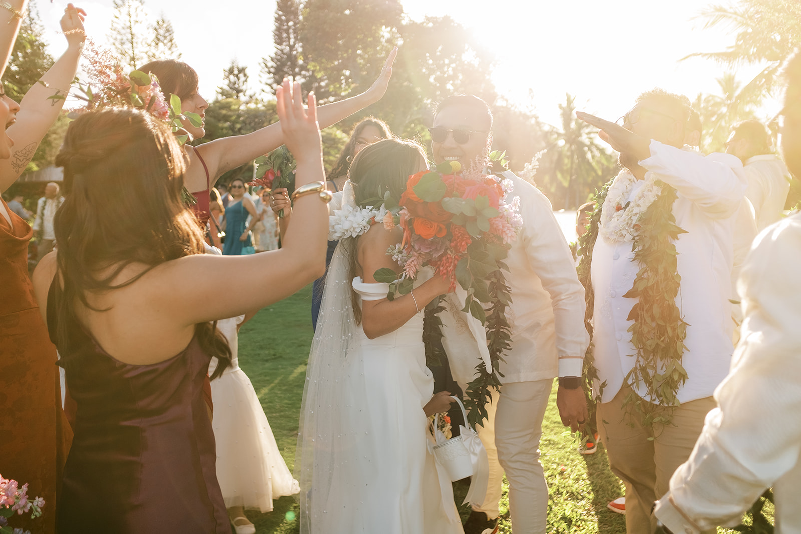 Newlyweds walking hand in hand as guests celebrate around them at an outdoor Hawaiian Wedding in Luau