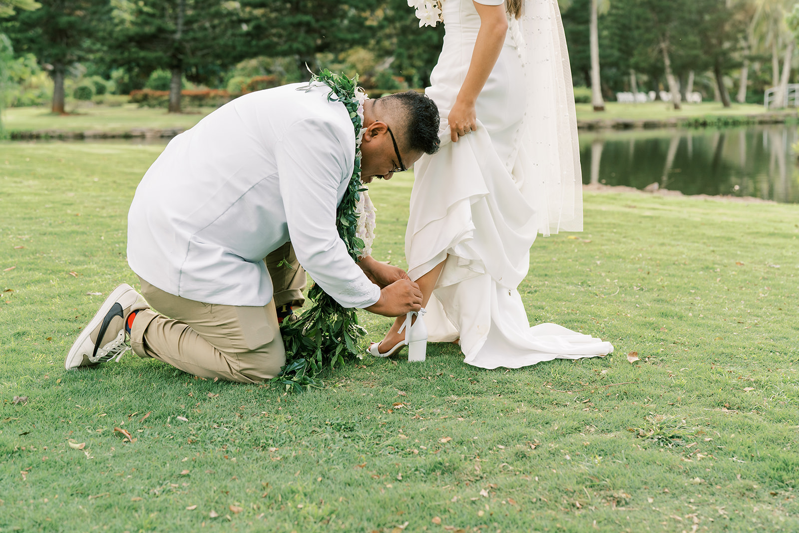 A groom adjusting the hem of a bride's dress outdoors.