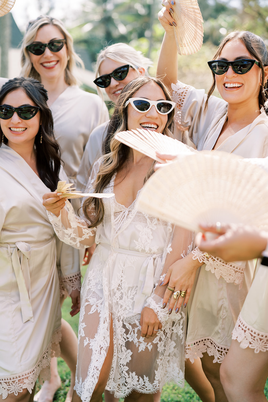 Women smiling and wearing sunglasses and matching robes, holding hand fans at a wedding in Kauai