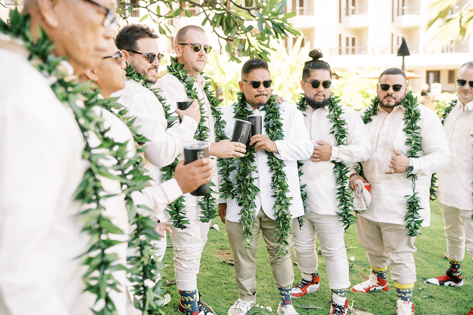 A group of men dressed in white shirts and beige pants with green leis, some wearing sunglasses, at wedding ceremony