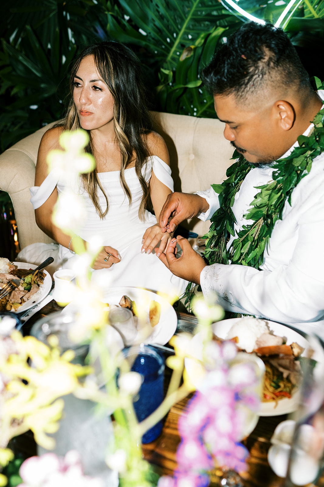 A bride sitting at a dinner table looks on as the groom, adorned with a lei, appears to be adjusting her wedding ring