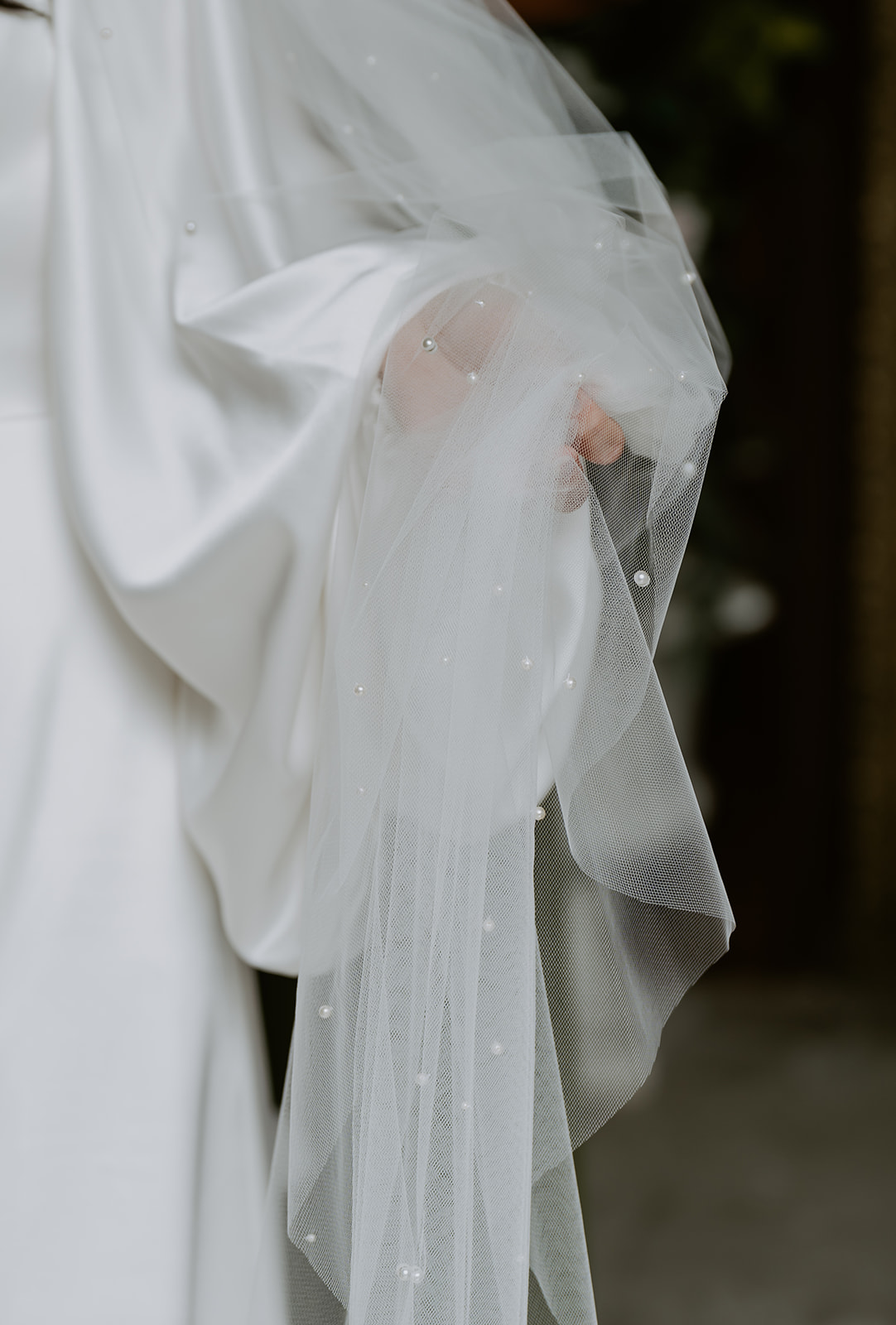 A bride adjusts her white veil adorned with pearls, her hand visible beneath the translucent fabric, with a natural back
