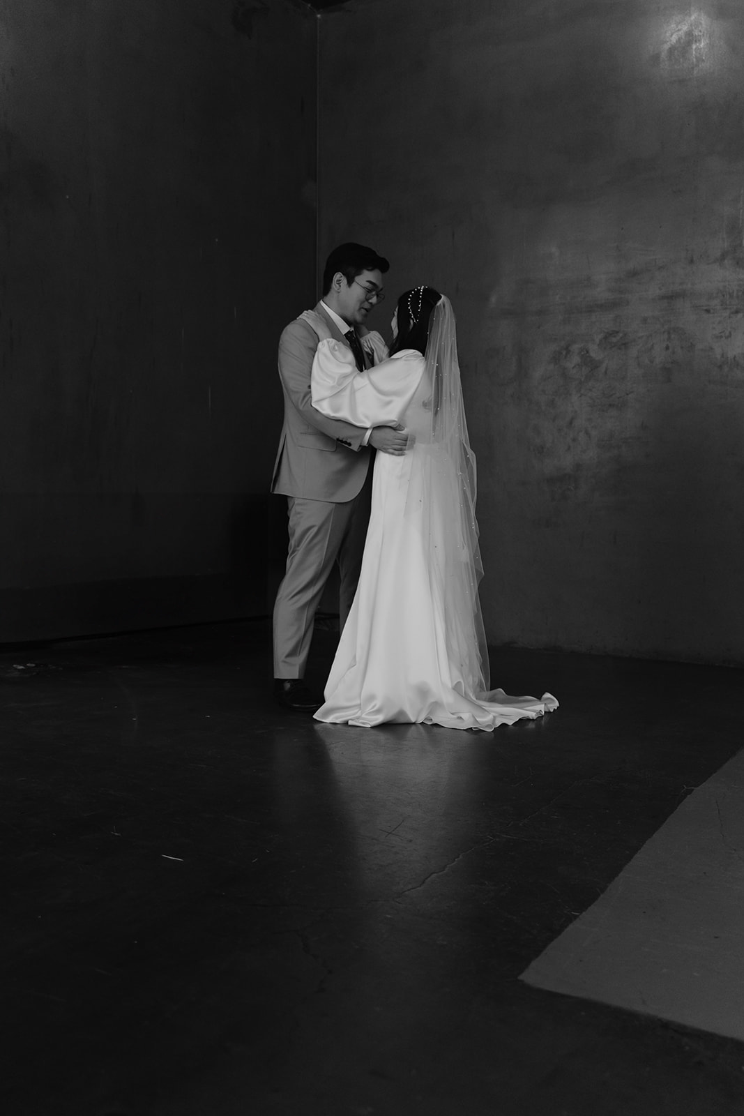 A bride and groom in elegant attire share a tender moment, dancing closely together in a dimly lit room.