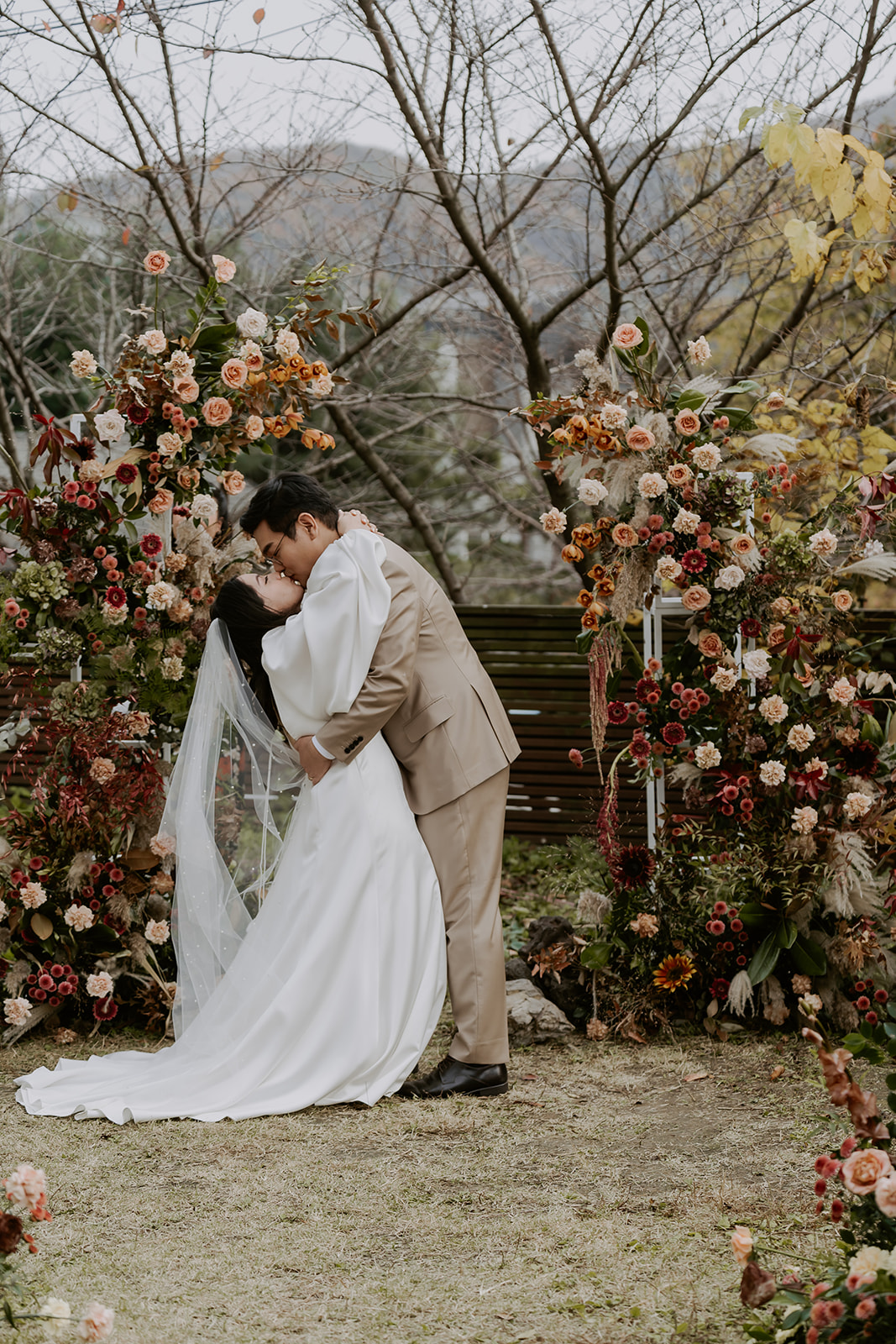 A bride and groom kissing under a floral arch in a garden setting, surrounded by colorful flowers and lush greenery.