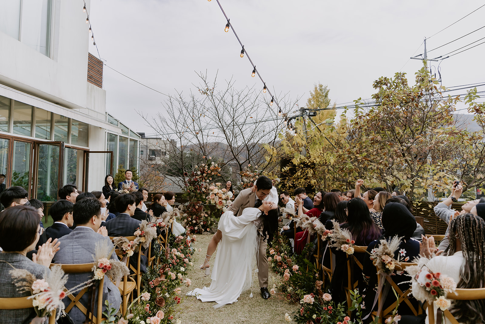 A bride and groom share a kiss on an aisle adorned with floral arrangements at an outdoor wedding ceremony with guests.