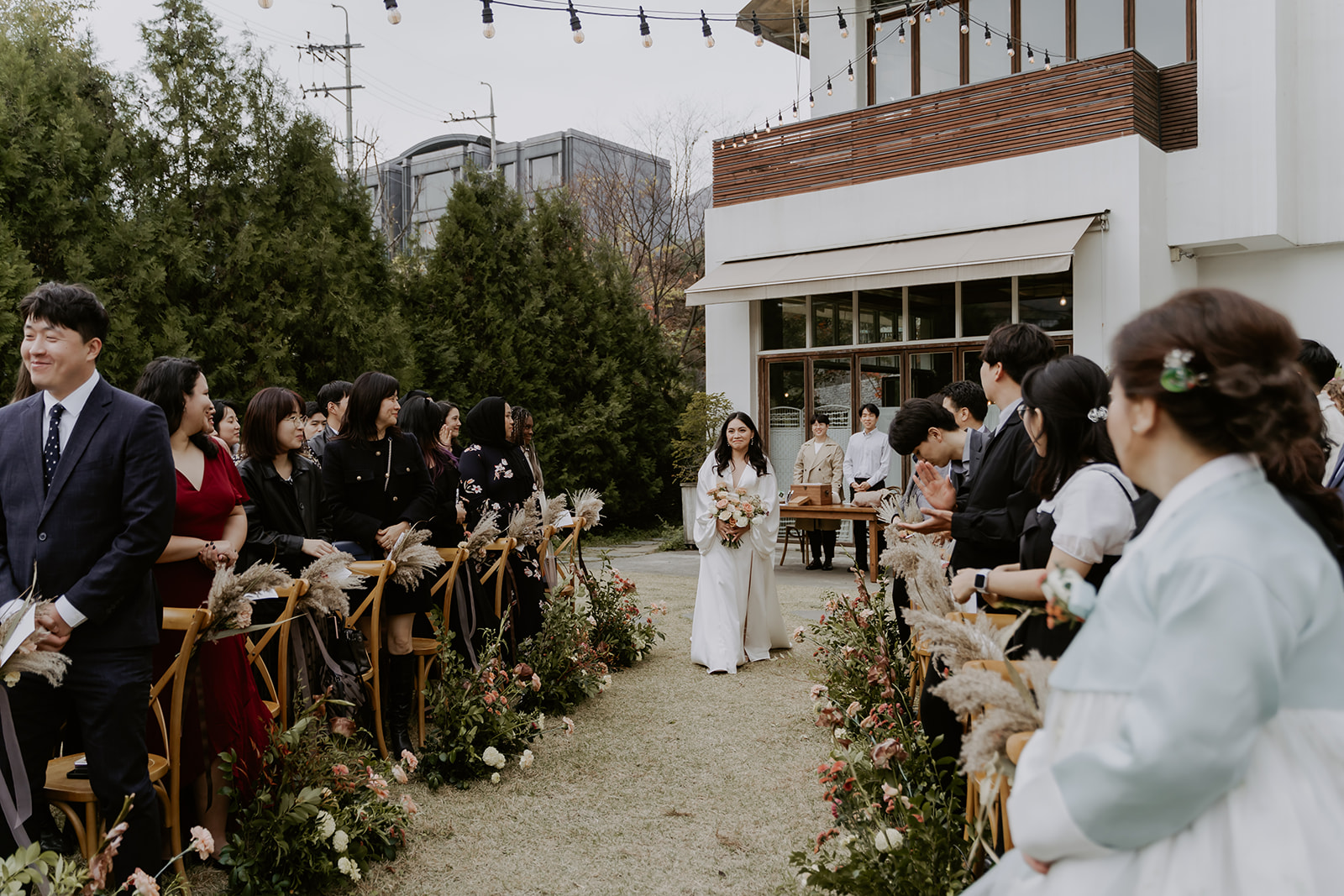 A bride walks down the aisle at an outdoor wedding ceremony, with guests lined up on either side, watching attentively.