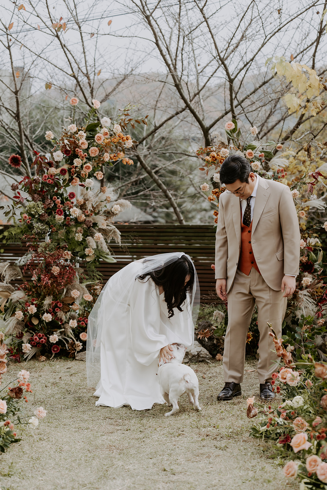 A bride in a white dress and a groom in a tan suit petting a small dog among floral decorations in a garden setting.