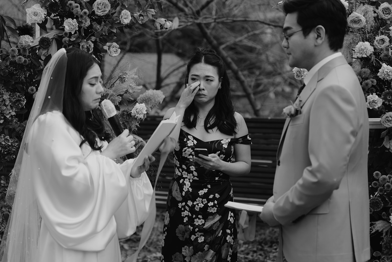 A bride in a white dress reads vows from a tablet at an outdoor ceremony, standing beside the groom in a suit.