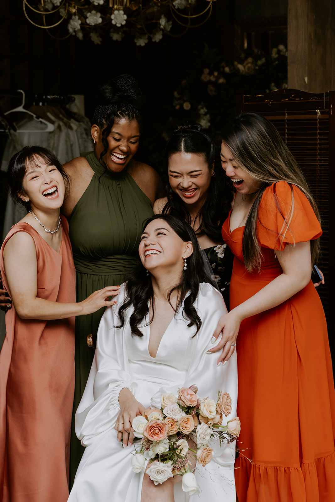 A bride in a white dress sits laughing with four bridesmaids in colorful dresses, holding a bouquet, in a rustic setting