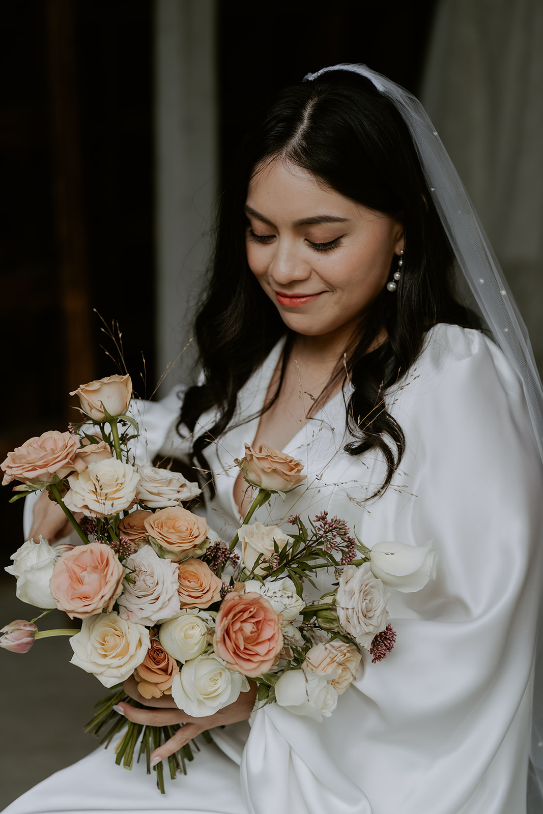 A bride in a white dress and veil smiling gently as she holds a bouquet of soft pink and cream roses.