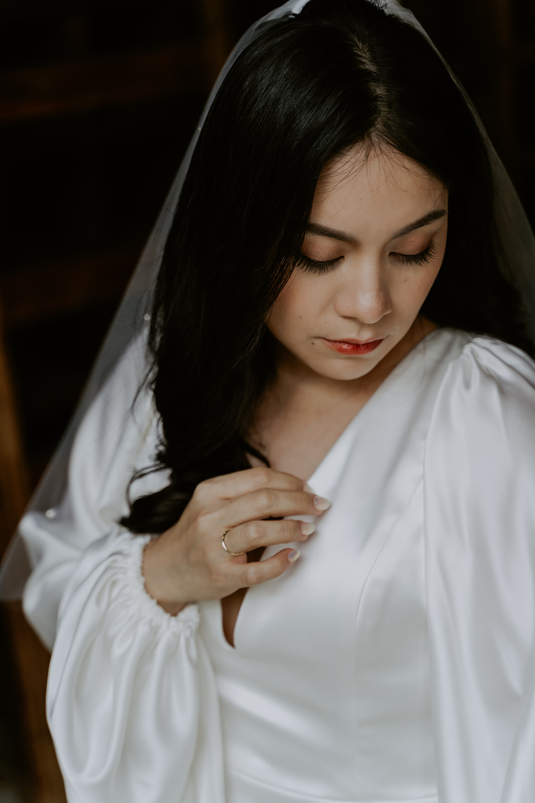 A bride in a white robe and veil, looking down while adjusting her necklace, showcasing a moment of quiet elegance.