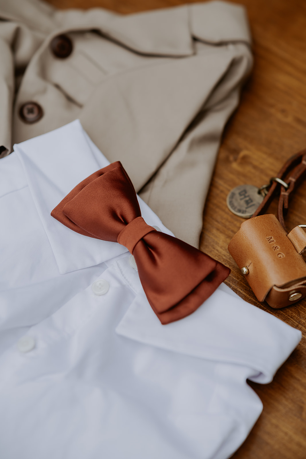 A brown bow tie rests on a white shirt with a beige jacket and tan leather accessories in the background.