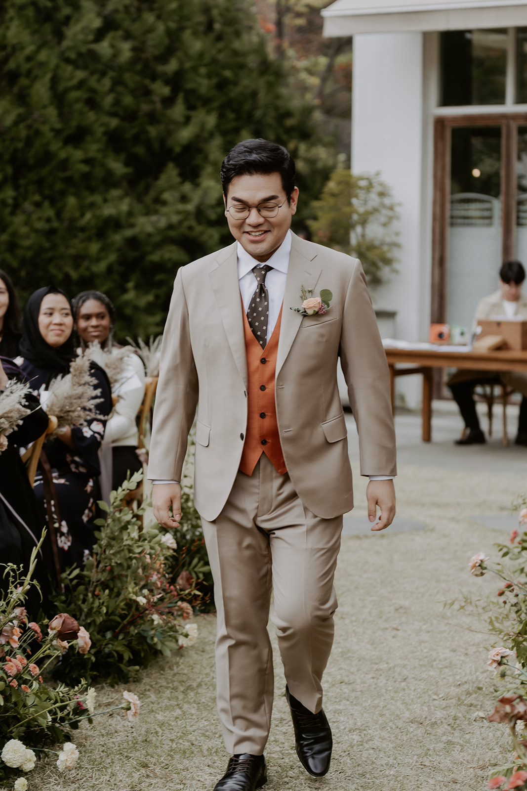 Man in a beige suit smiling as he walks down a garden aisle at a wedding, with seated guests in the background.