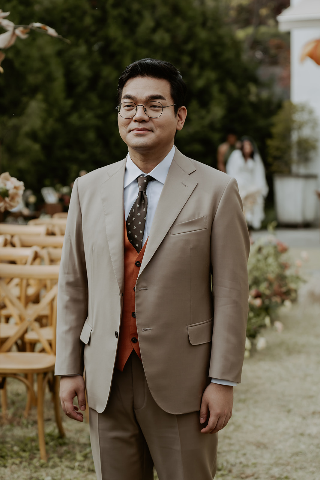 Man in elegant suit smiling at an outdoor wedding ceremony, with a bride visible in the background.