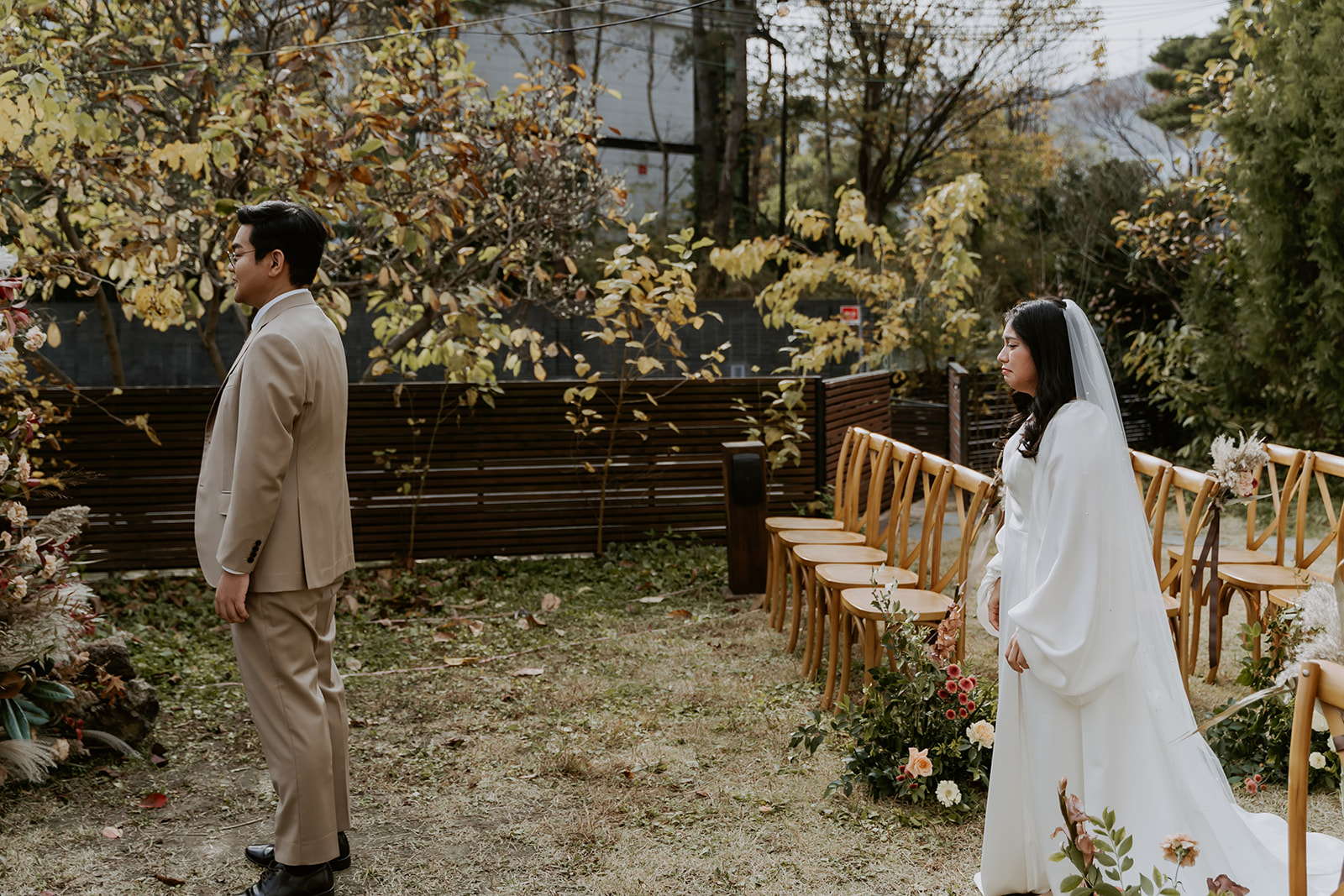 A man and woman in formal attire at their first look in a garden with autumn foliage.