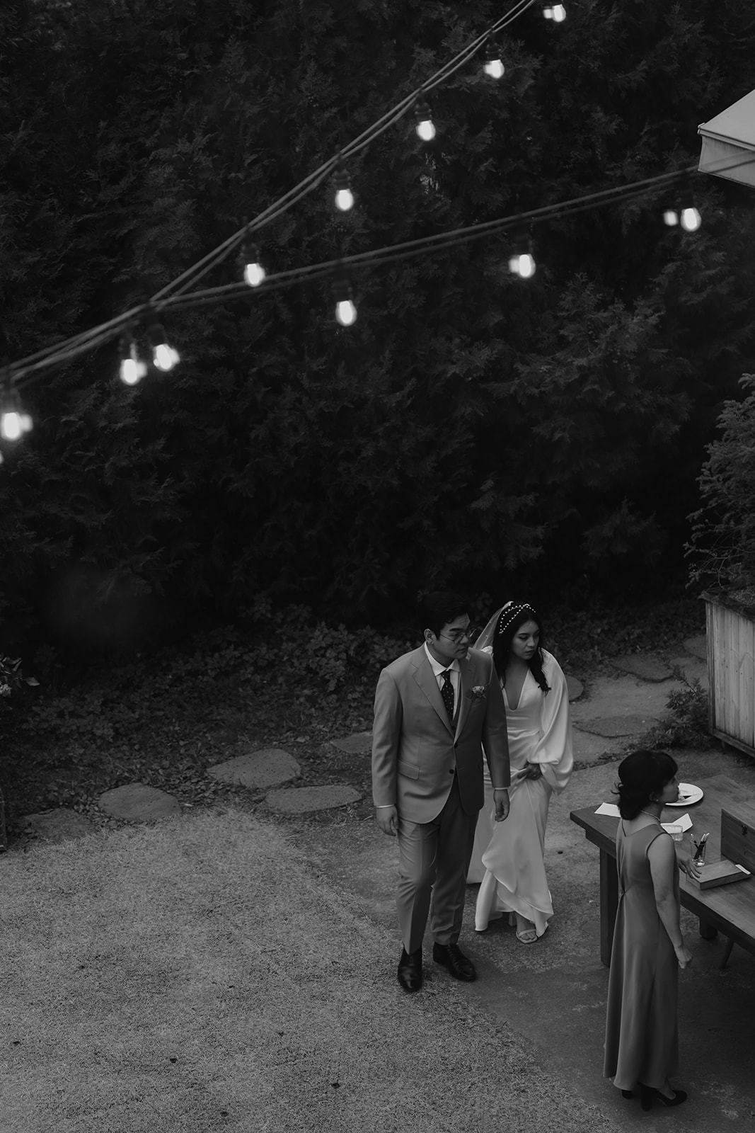 a newlywed couple walking hand in hand at an outdoor venue, greeted by a guest under string lights.