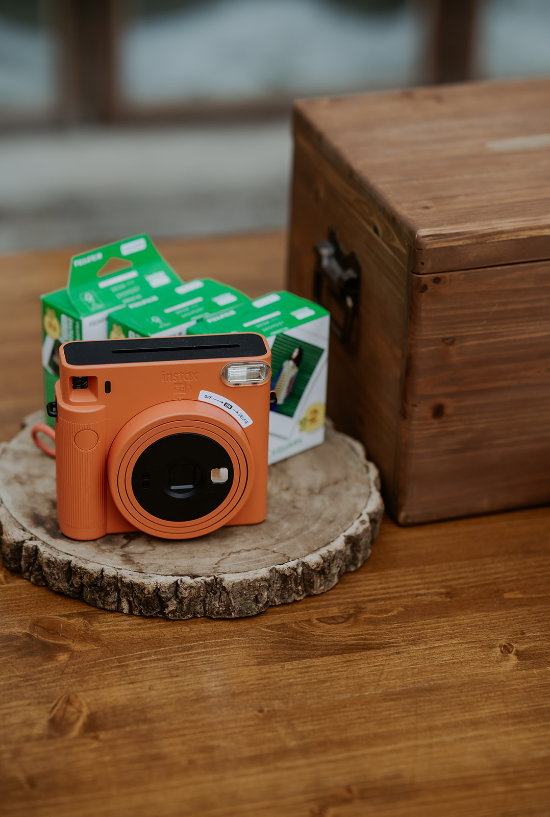 An orange fujifilm instax camera and film boxes placed on a wooden slice next to a closed wooden box.