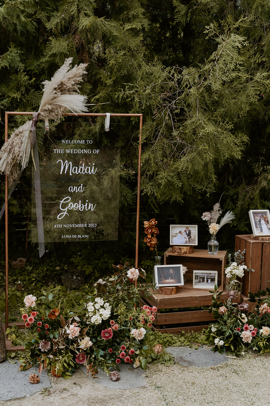 A rustic wedding welcome sign with the names "madai and gobin" surrounded by flowers and framed pictures.