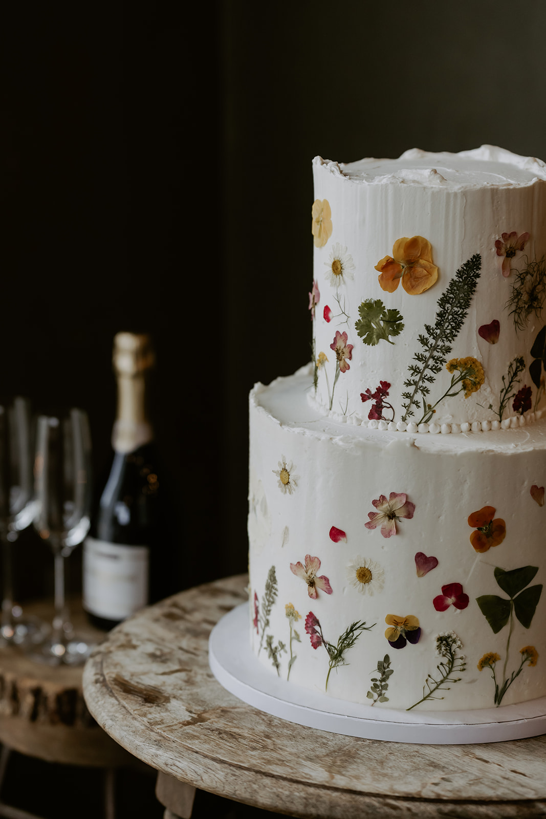 A tiered white cake adorned with edible flowers, displayed on a rustic table next to champagne flutes and a bottle.