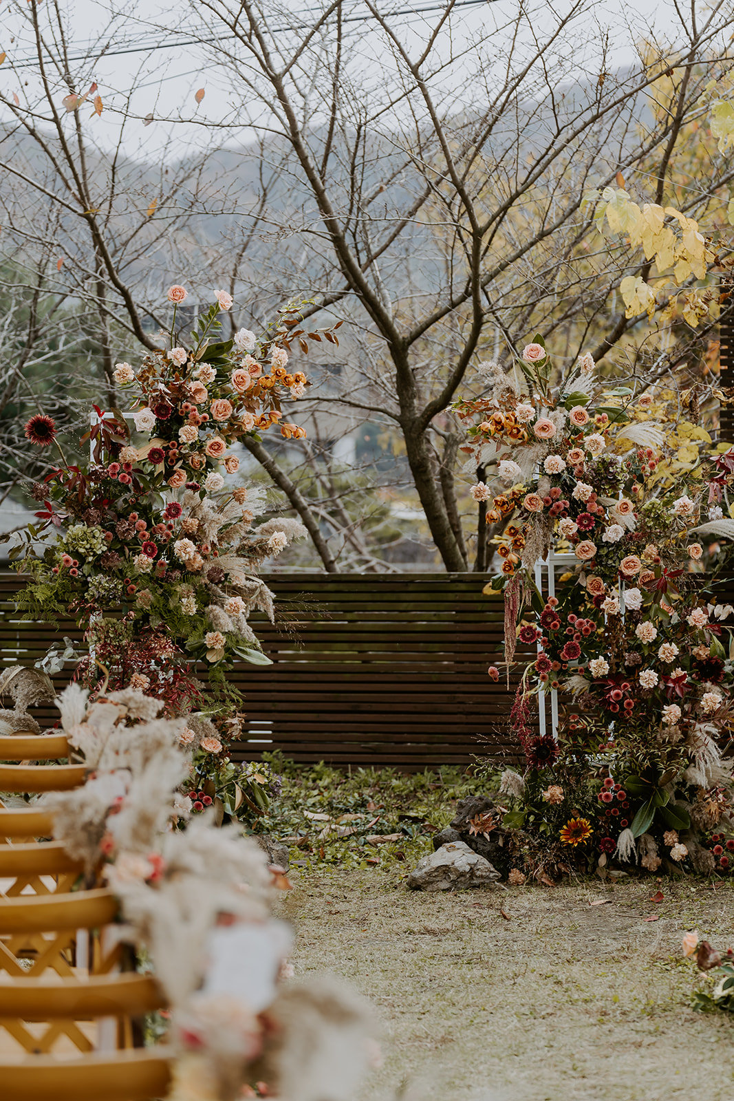 Two large floral arches adorn an outdoor wedding aisle, set against a natural backdrop with autumn foliage and a rustic 
