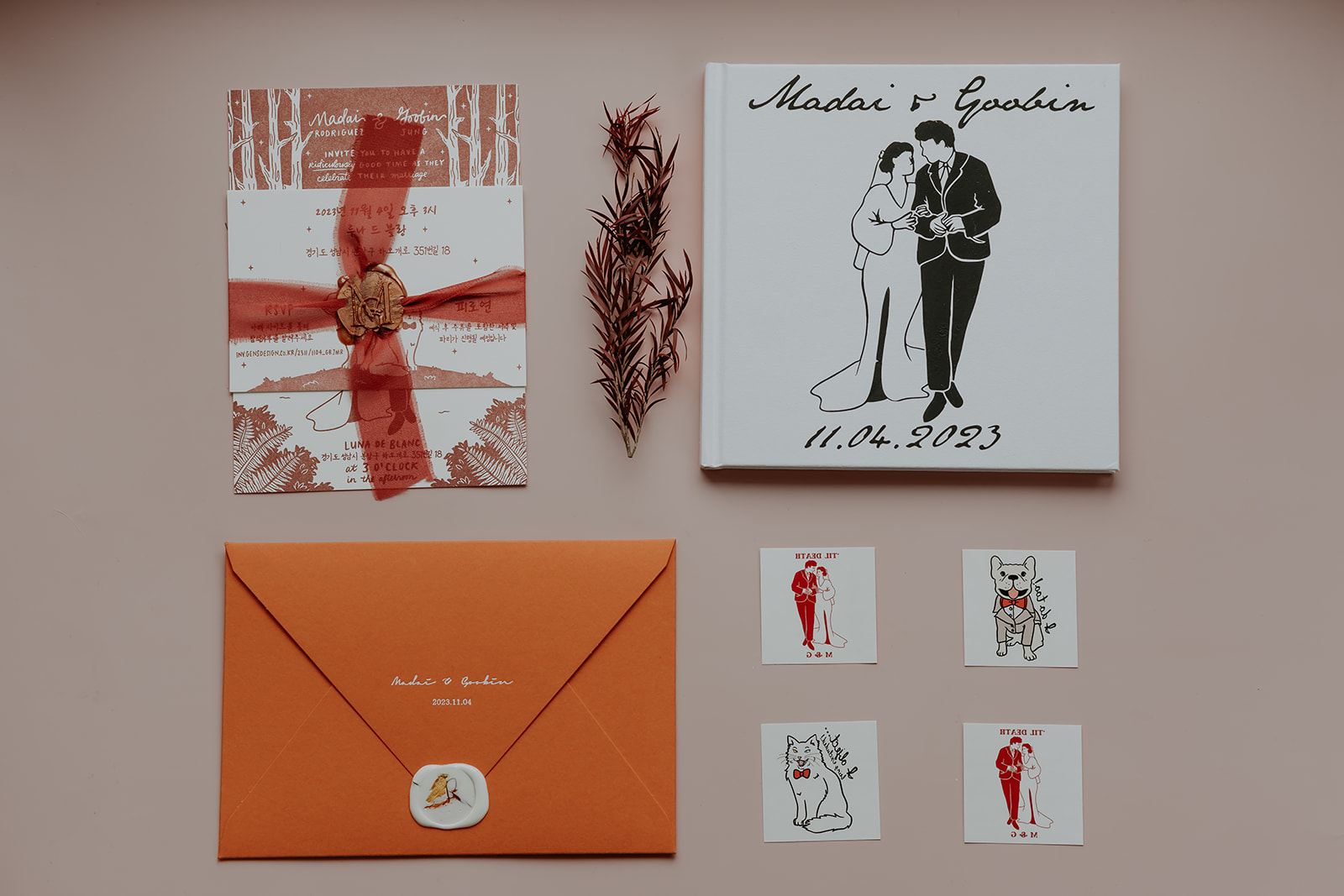 Wedding invitation suite featuring an orange envelope, invitation card with red wax seal, guest book, and small cartoon 