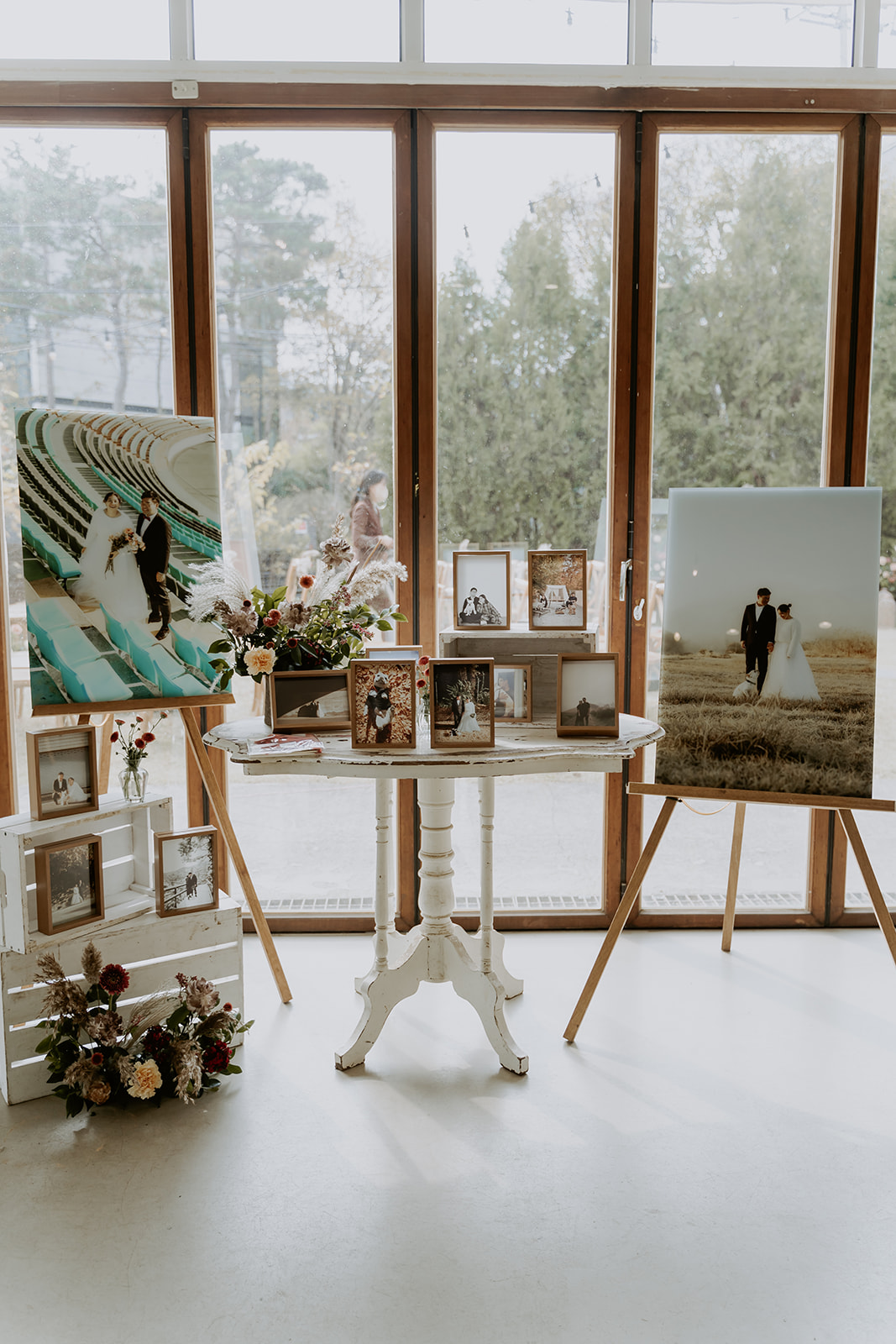A wedding photo display featuring frames on a shelf, easels, and a table with floral decorations in a room.