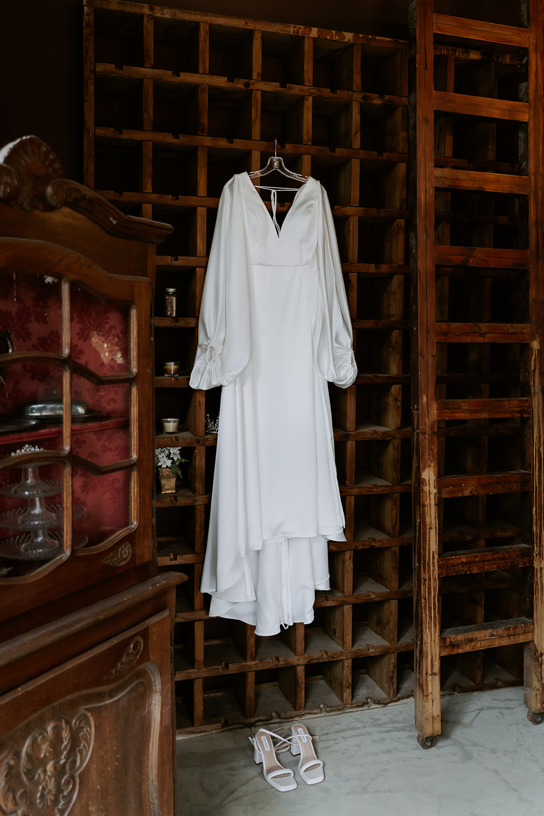A white wedding dress and a pair of shoes hanging in front of a large wooden shelf with cubbies filled with various item