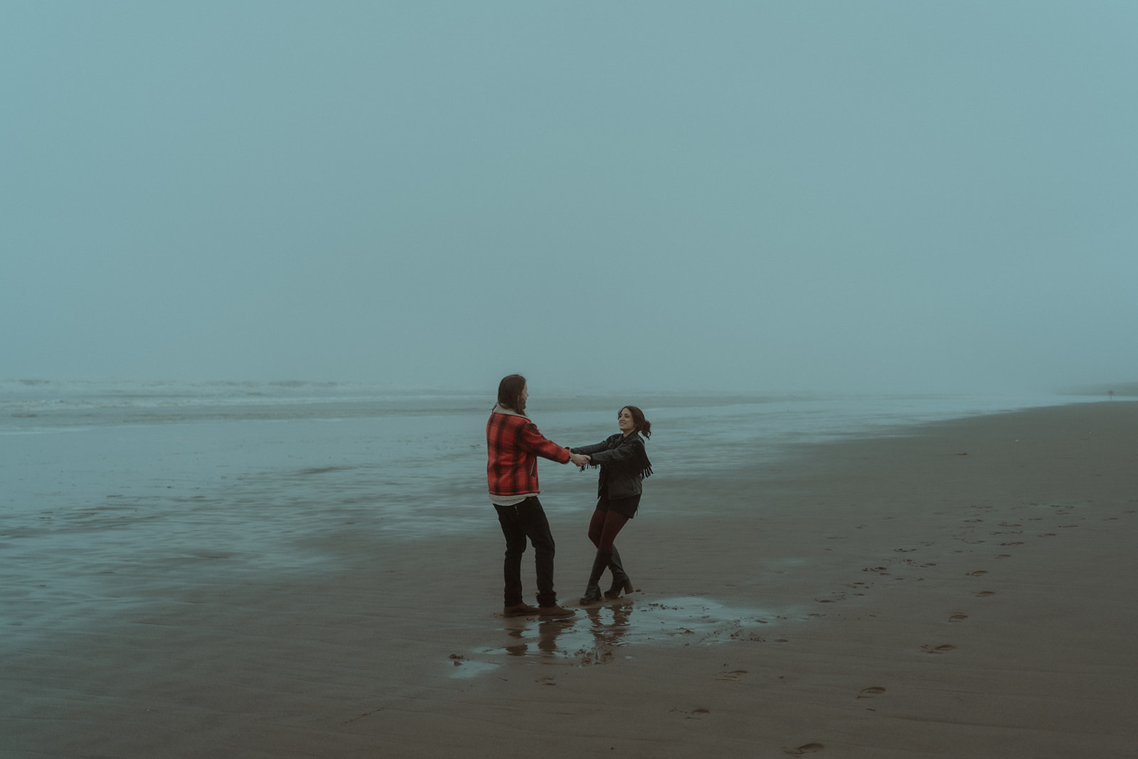 Documentary-style PNW Oregon coast engagement photo of couple holding hands and spinning on the beach