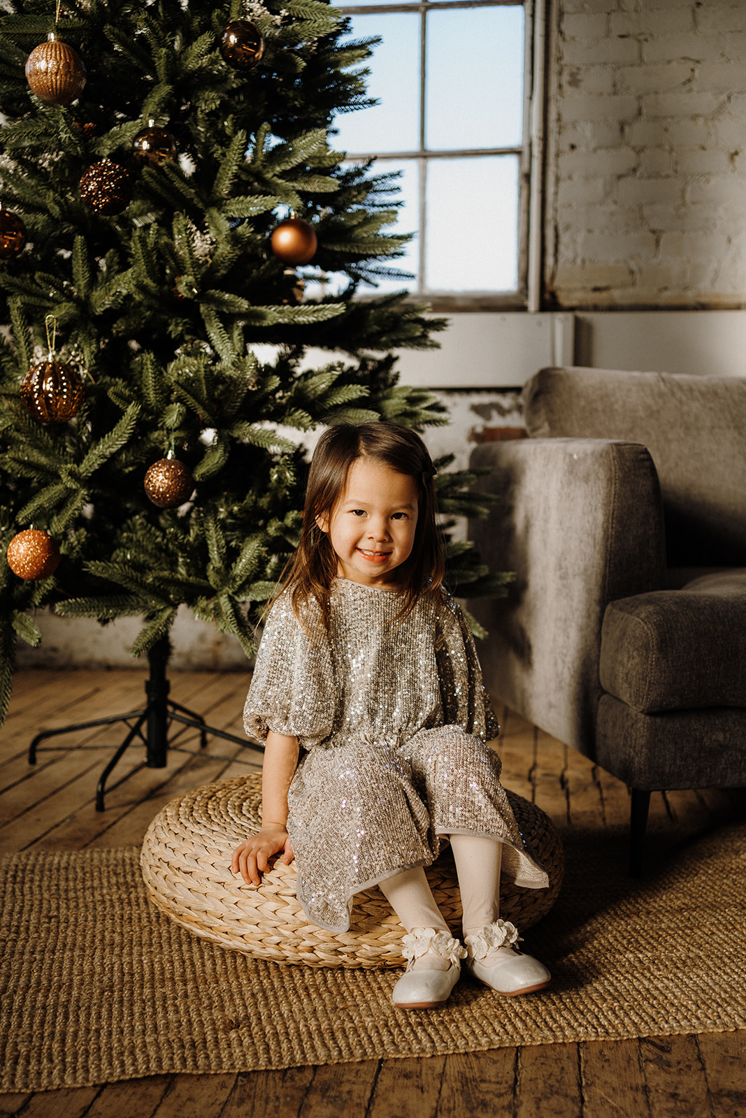 A little girl sitting on a cushion on the ground in front of the Christmas Tree.