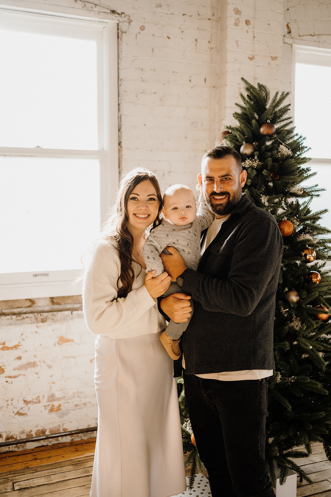 Family of three standing inside in front of a Christmas Tree.