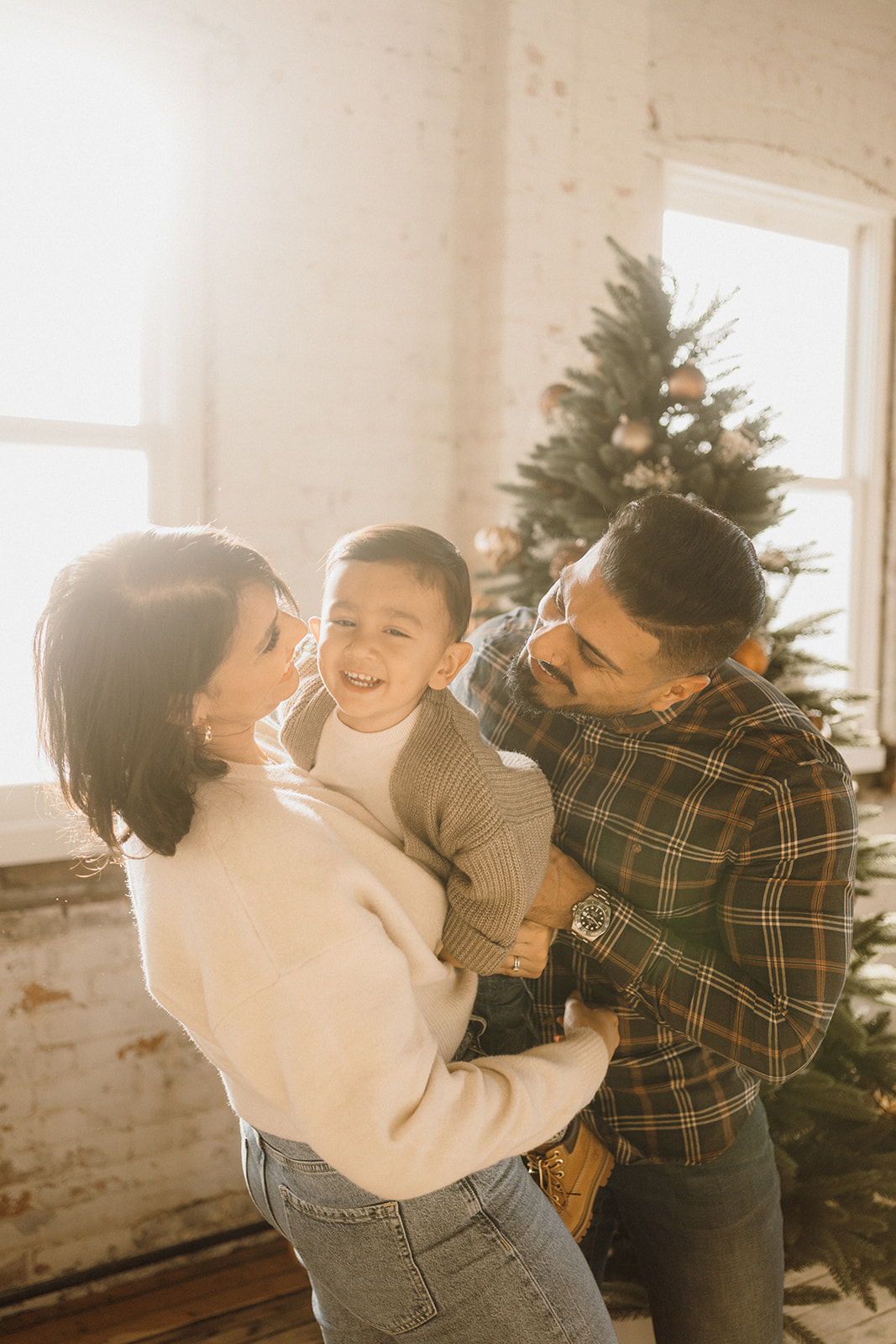 A family of three inside in front of a Christmas Tree.