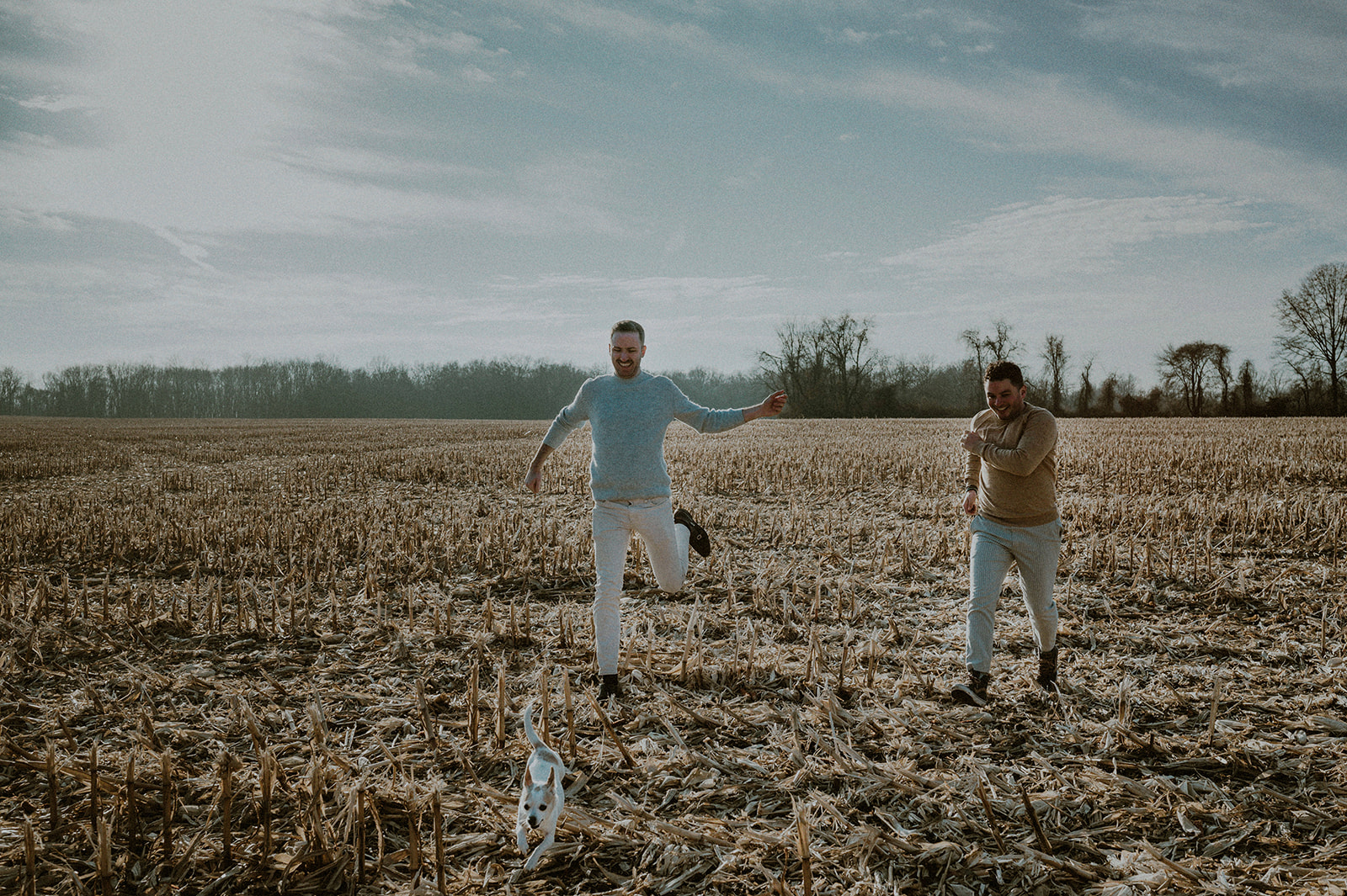 two men running in bare NJ corn field with chihuahua during their engagement session