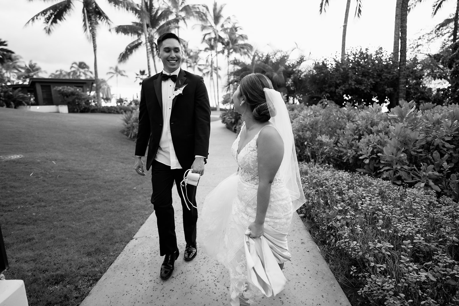 A black and white photo of a bride and groom walking down a path in front of palm trees at the Four Seasons Oahu