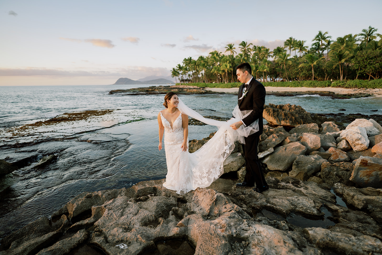 Groom holding the trail of the bride's wedding dress at sunset on The Four Seasons Resort Oahu