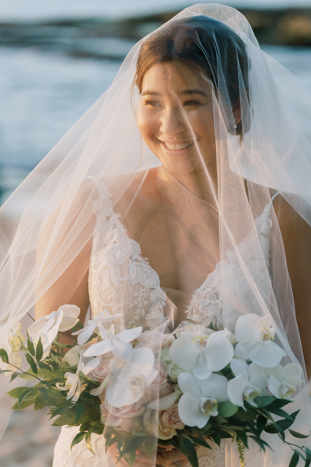 A bride smiles while holding her wedding bouquet on the beach.