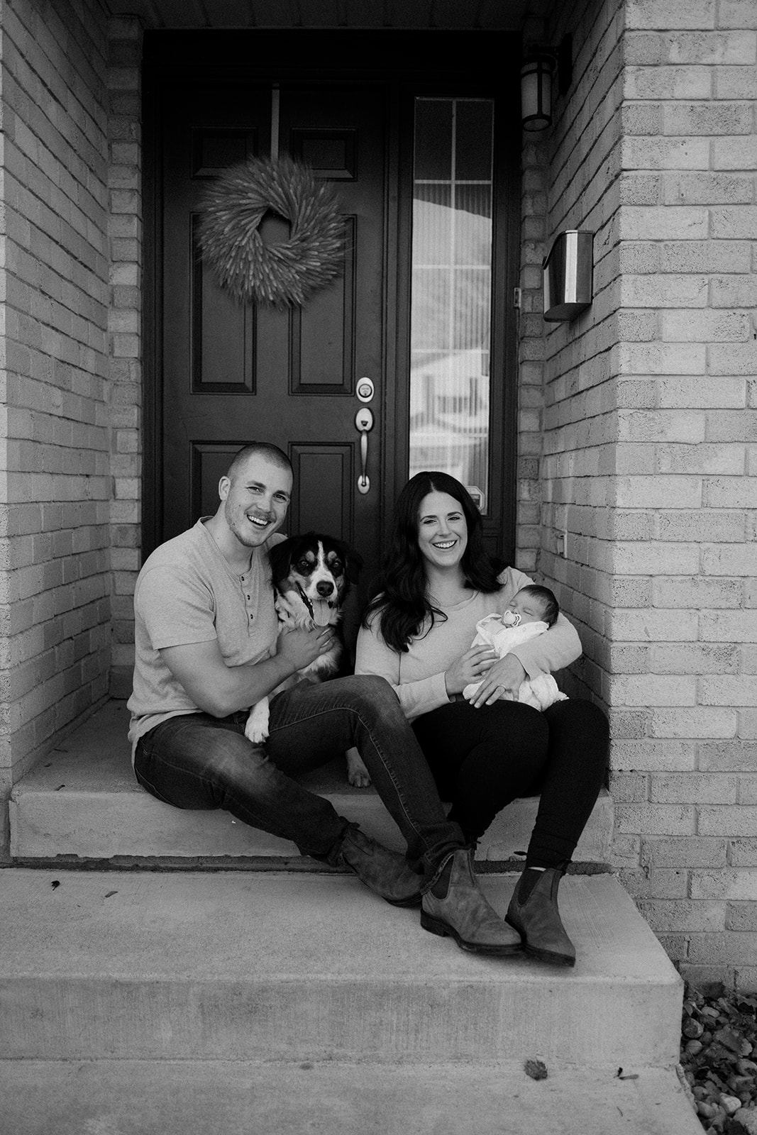 Parents sitting on their porch outside with their newborn and puppy beside them.