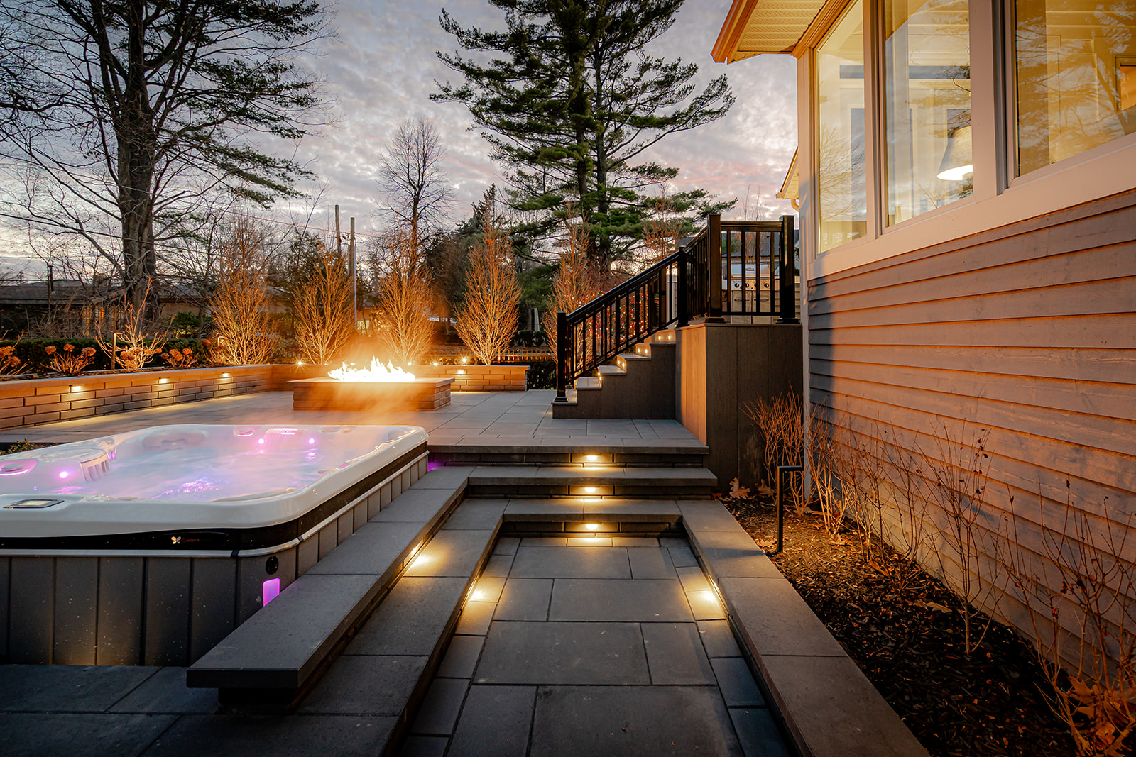 A jacuzzi with lights on and a fireplace with stairs leading to the house.