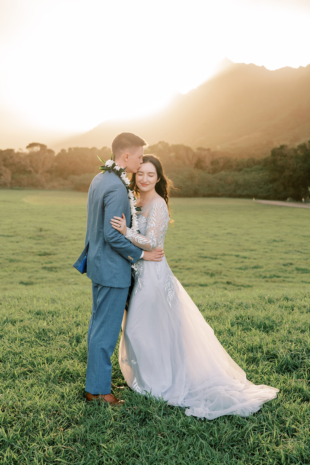 A bride and groom embrace in a field at Kualoa Ranch during their destination wedding at sunset.