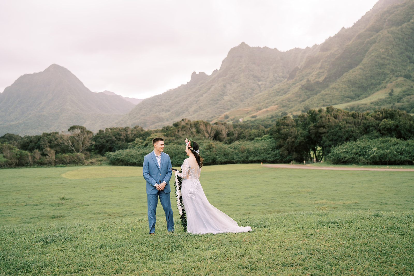 A bride and groom standing in a field at Kualoa Ranch with mountains in the background.