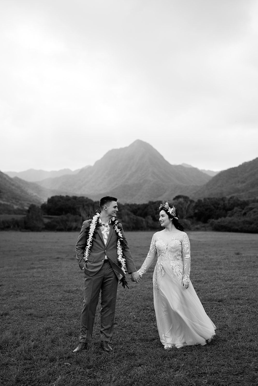 A monochorome photo of the bride and groom standing in a field at Kualoa Ranch with mountains in the background.