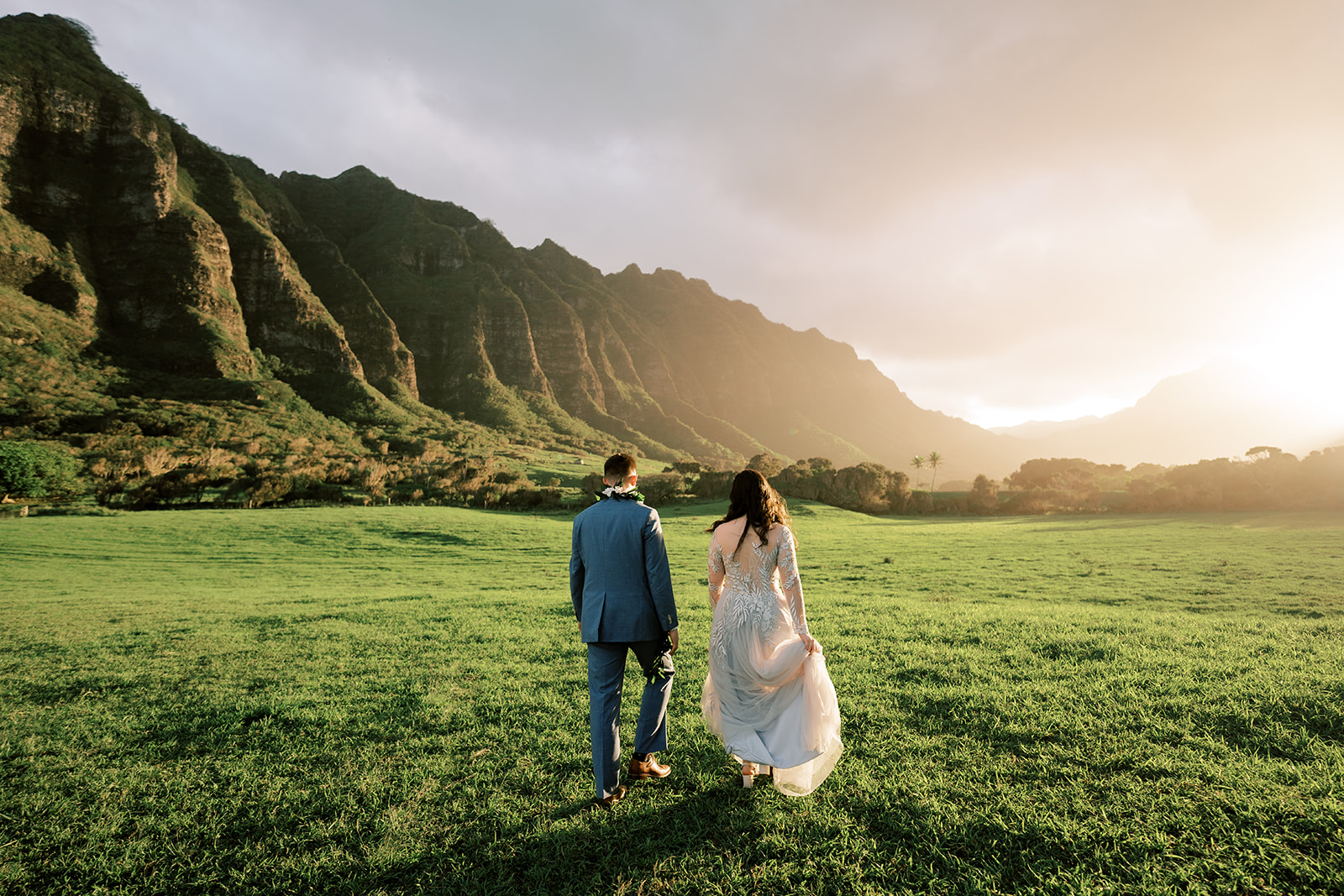 A bride and groom walking through a grassy field at sunset.